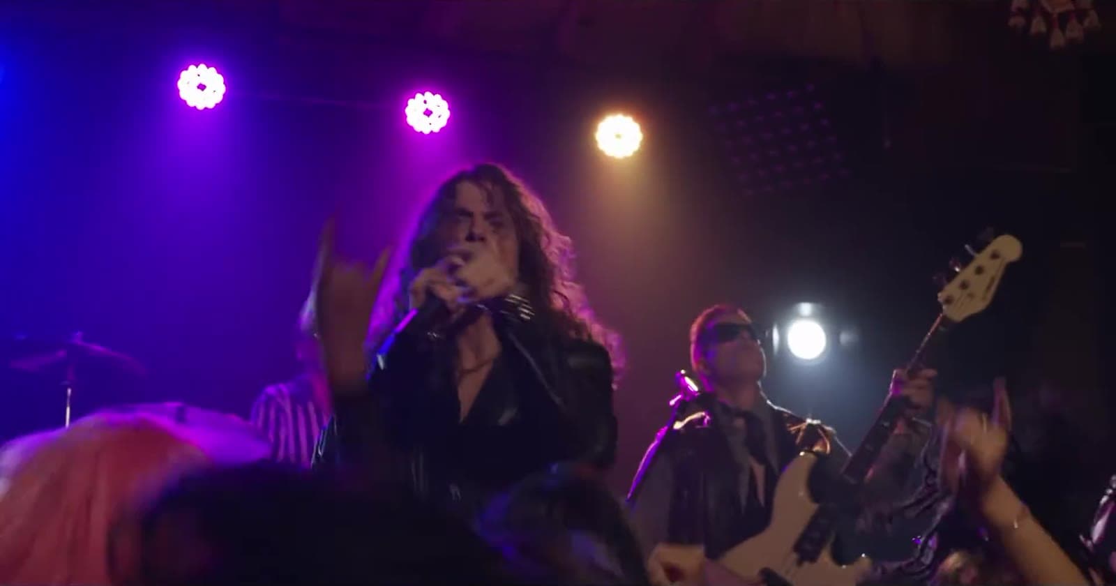 The singer at the microphone with the bassist and stage lights behind, and the crowd's hands in the foreground